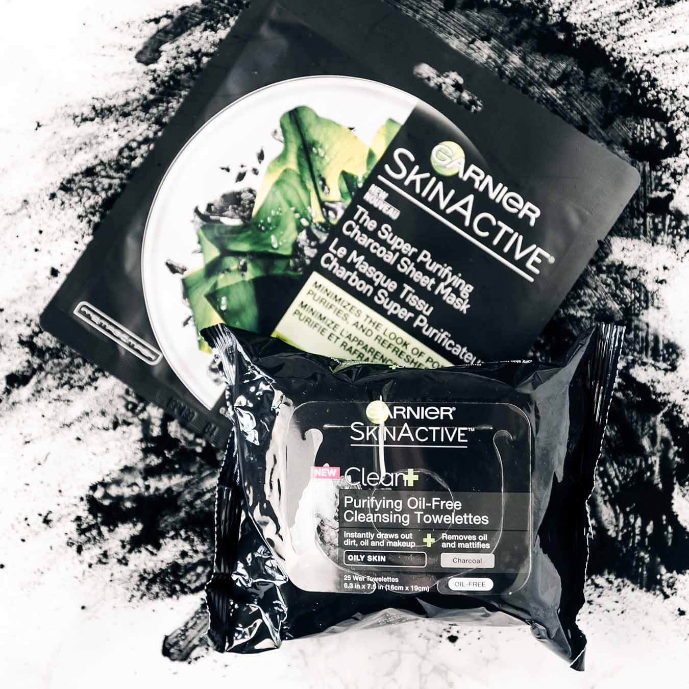 Garnier SkinActive Super Purifying Charcoal Sheet Mask and SkinActive Clean+ Purifying Oil-Free Cleansing Towelettes on an explosion of charcoal powder on pinkish white marble.